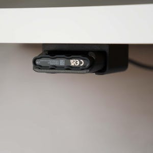 May include: A black under-desk mount holding a black external hard drive. The drive features a textured edge, a QR code, and printed text including 'UK CA' and 'CE'. The mount is secured to the underside of a white surface, providing a storage solution.