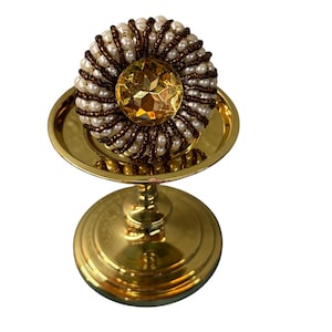 May include: A gold-coloured decorative stand with a round top and a pedestal base. The top features a large, faceted, yellow gemstone surrounded by white pearls and brown beads. The stand is likely used for display.