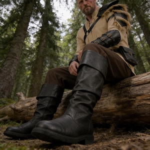 Puede incluir: Botas negras de cuero hasta la rodilla con puño doblado. Las botas tienen un aspecto texturizado y una suela resistente. La imagen muestra a una persona que lleva las botas, junto con una camisa color canela, pantalones marrones y una pieza de hombro de cuero, en un entorno forestal.