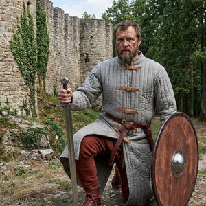 Könnte beinhalten: Mittelalterliche Kleidung mit einer grauen, gesteppten Tunika mit Lederriemen, einer rotbraunen Hose und einem braunen Ledergürtel. Das Ensemble umfasst einen Holzschild und ein Schwert. Im Hintergrund eine Steinmauer.