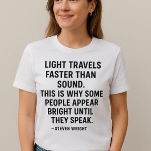 May include: White t-shirt with the quote "LIGHT TRAVELS FASTER THAN SOUND. THIS IS WHY SOME PEOPLE APPEAR BRIGHT UNTIL THEY SPEAK. - STEVEN WRIGHT" in black text. The shirt is worn by a person with brown hair and blue jeans.