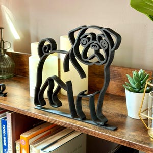 以下が含まれることがあります： 棚に置かれた、黒いレーザーカットのパグ犬のシルエット。装飾品は、丸まった尾や特徴的な顔など、犬の特徴の詳細な輪郭を示しています。パグの高さは約20cmです。