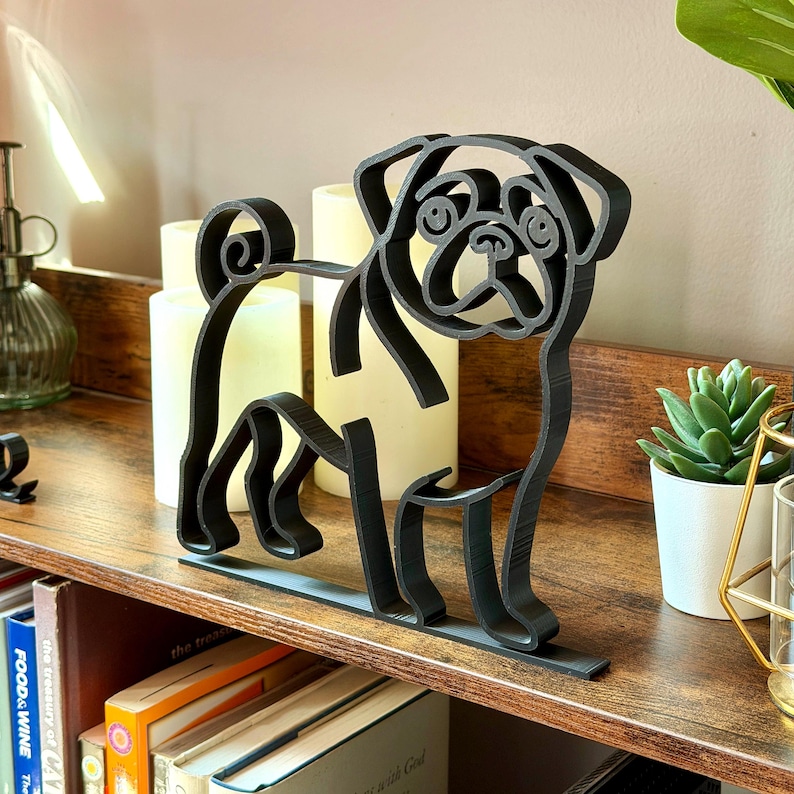 Minimalist pug line art sculpture in matte black shown on a wooden bookshelf with candles, books, and plants. This modern dog shelf sitter decor captures the pug’s playful face, curly tail, and sturdy stance. Perfect as bookshelf decor, reading nook art, or a gift for pug lovers. Lightweight 3D printed sculpture blends with modern, Scandinavian, and eclectic home styles. Ideal office decor or pet lover gift.