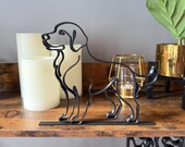 Minimalist black line art dog sculpture, 3D printed in matte black filament from a single continuous line. Depicts a standing Labrador or Golden Retriever with floppy ears, friendly face, and curved tail. Mounted on a flat base, displayed on a rustic wooden shelf with LED candles, gold candle holder, and potted plant in gold pot. Perfect modern decor for dog lovers, pet memorials, or contemporary home accents.