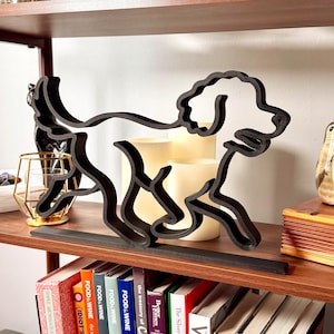 May include: A black, stylized wooden dog silhouette, possibly a poodle, is displayed on a shelf. The dog is running, with its legs and tail extended. The shelf also holds books, candles, and decorative objects.