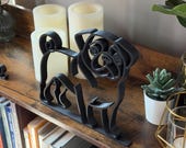 Angled photo of pug line art shelf sitter sculpture styled on a bookshelf with greenery, books, and candles. The smooth black outline captures the pug’s playful charm in a modern minimalist way. Perfect shelf decor for reading nooks, offices, or living rooms. Lightweight 3D printed sculpture makes a thoughtful dog lover gift. Versatile art piece for housewarming, birthday, or holiday presents.