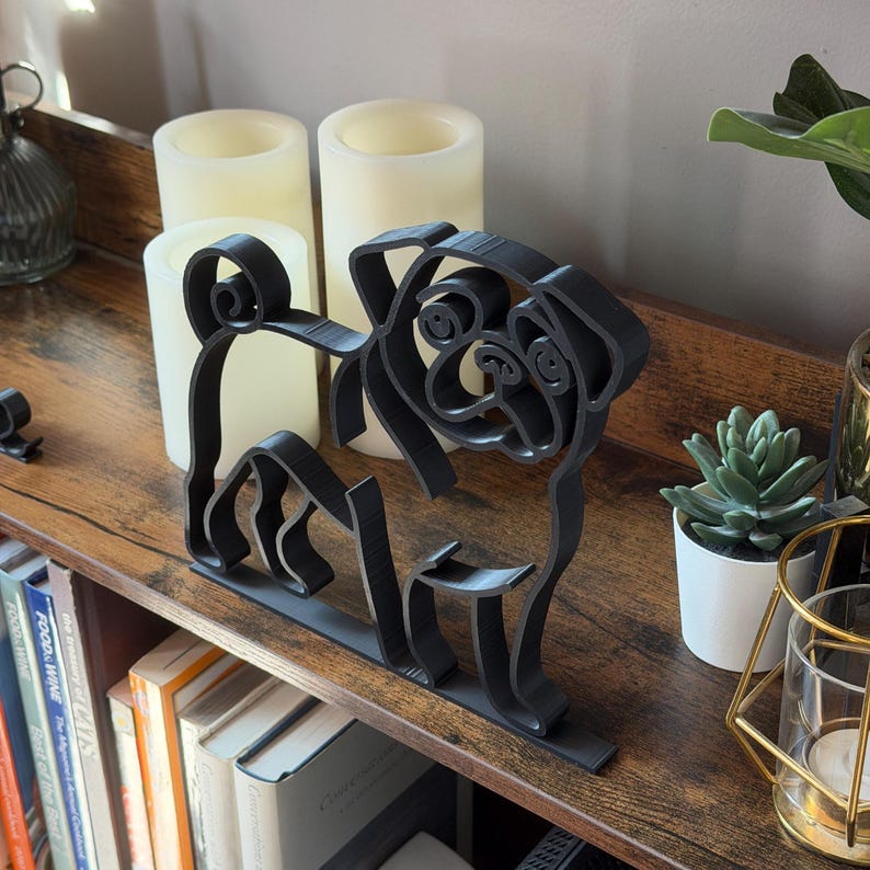 Angled photo of pug line art shelf sitter sculpture styled on a bookshelf with greenery, books, and candles. The smooth black outline captures the pug’s playful charm in a modern minimalist way. Perfect shelf decor for reading nooks, offices, or living rooms. Lightweight 3D printed sculpture makes a thoughtful dog lover gift. Versatile art piece for housewarming, birthday, or holiday presents.
