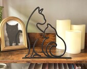 Minimalist black 3D printed cat line art sculpture featuring two cats sitting together, displayed on a rustic wood shelf with candles and books in the background. The sleek modern design adds a cozy, artistic touch to cat-themed home decor. Ideal for cat lovers, boho interiors, or as a thoughtful handmade gift.
