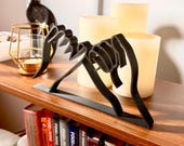 Modern 3D printed pinky promise sculpture made from black minimalist line art, displayed on a wooden shelf. This handmade line art decor features two hands linked in a pinky swear, symbolizing trust, loyalty, and love. Ideal as a friendship gift, romantic anniversary decoration, or stylish addition to modern and boho home decor. Surrounded by white candles and gold accents, the sculpture creates a warm, aesthetic vibe.