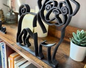 Close-up of pug minimalist sculpture displayed on a bookshelf beside candles and a potted plant. The sleek black line art design highlights the pug’s wrinkled face, curled tail, and compact body. Unique bookshelf decor for dog lovers, modern office accents, or living room shelf styling. 3D printed design adds personality and charm to any room. Perfect gift idea for pug enthusiasts, birthdays, or housewarming.