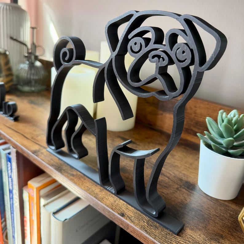 Close-up of pug minimalist sculpture displayed on a bookshelf beside candles and a potted plant. The sleek black line art design highlights the pug’s wrinkled face, curled tail, and compact body. Unique bookshelf decor for dog lovers, modern office accents, or living room shelf styling. 3D printed design adds personality and charm to any room. Perfect gift idea for pug enthusiasts, birthdays, or housewarming.