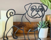 Straight-on view of pug line art sculpture on wooden shelf with candles and plants. Matte black minimalist design highlights pug’s wrinkled face, curly tail, and small sturdy frame. Perfect as bookshelf decor, office accents, or modern shelf styling. Unique handmade look without mass-production feel. 3D printed sculpture makes an ideal pug lover gift for birthdays, holidays, or housewarming.