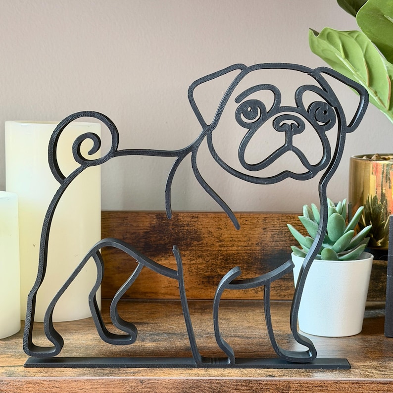 Straight-on view of pug line art sculpture on wooden shelf with candles and plants. Matte black minimalist design highlights pug’s wrinkled face, curly tail, and small sturdy frame. Perfect as bookshelf decor, office accents, or modern shelf styling. Unique handmade look without mass-production feel. 3D printed sculpture makes an ideal pug lover gift for birthdays, holidays, or housewarming.