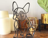 Minimalist French Bulldog line art sculpture in black PLA, sitting on a wooden shelf with candles and gold decor in the background.