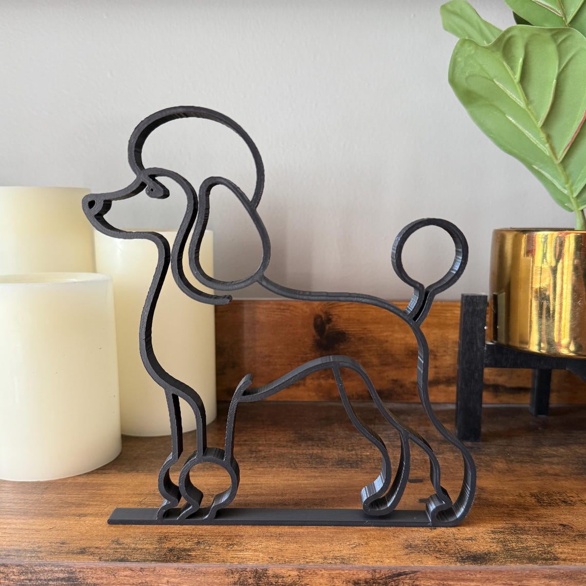Minimalist black poodle line art sculpture in PLA plastic, standing on a wooden shelf with candles and a gold planter in the background.