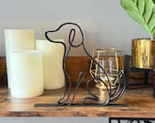 A full-frontal view of a black minimalist dog sculpture made from a single looping line, 3D printed in matte filament and displayed on a wooden shelf. The abstract silhouette of the sitting dog is elegant and modern, with a simple flat base and no floating elements. Surrounded by creamy LED candles, a gold candle holder, and a small potted plant, this pet-themed decor is ideal for dog lovers seeking a unique shelf accent, office desk ornament, or housewarming gift.