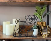 Modern pug sculpture in continuous line art style, matte black finish, set on a rustic wood shelf with candles and decor. Captures the iconic pug expression and curly tail in a sleek, artistic silhouette. Makes stylish bookshelf decor, desk accent, or gift for pug owners. Lightweight and durable design fits with minimalist, eclectic, or Scandinavian home interiors. Ideal shelf sitter sculpture for dog lovers.