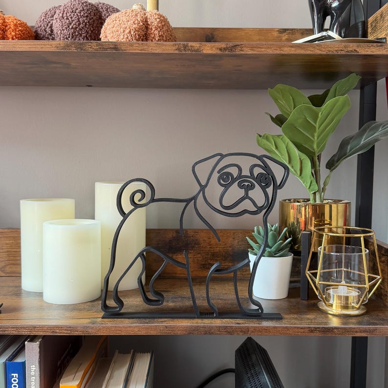 Modern pug sculpture in continuous line art style, matte black finish, set on a rustic wood shelf with candles and decor. Captures the iconic pug expression and curly tail in a sleek, artistic silhouette. Makes stylish bookshelf decor, desk accent, or gift for pug owners. Lightweight and durable design fits with minimalist, eclectic, or Scandinavian home interiors. Ideal shelf sitter sculpture for dog lovers.