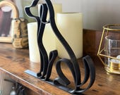 A side view of a modern abstract dog sculpture crafted from one continuous black line, 3D printed with visible layers and designed for a clean, minimalist aesthetic. The figure is shaped into a sitting dog with a curved tail and upright ears, attached to a flat black base. Set on a rustic wooden shelf with soft LED candles and gold accents, this decorative piece blends contemporary design with pet-inspired art, perfect for dog lovers, pet memorials, or entryway styling.