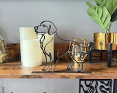 Sleek black Labrador or Golden Retriever sculpture, created from one continuous line and 3D printed in matte black. Depicts floppy ears, friendly face, and poised stance with tail. Displayed on a rustic wood shelf with LED candles, gold candle holder, and plant in gold pot. Great for dog lovers, pet memorials, or modern home decor accents.