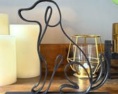 A minimalist black line art dog sculpture made from a single continuous line, 3D printed in matte black filament and placed on a natural wood shelf. The decor features a stylized silhouette of a sitting dog with smooth, curved lines and a flat base for stability. It’s styled next to neutral-toned LED pillar candles and a gold geometric candle holder, making it an ideal modern home accent for dog lovers and contemporary decor themes.