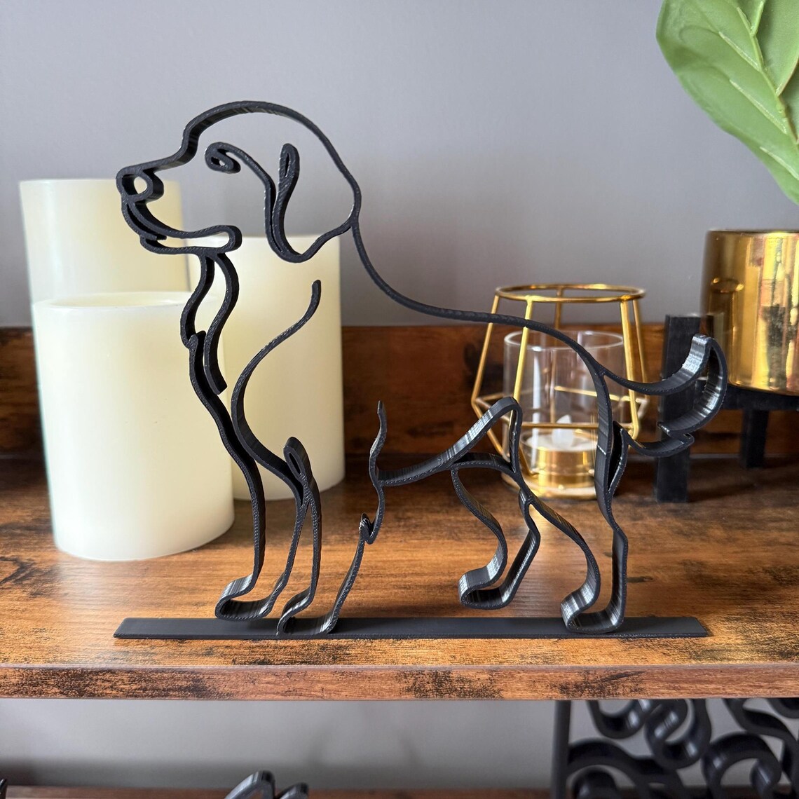 Front view of a modern Labrador or Golden Retriever line art sculpture, 3D printed in matte black from one flowing line. Features floppy ears, alert stance, and wagging tail on a flat black base. Displayed on a wood shelf with LED candles, gold candle holder, and greenery in gold pot. Ideal for dog-themed gifts, pet memorials, or stylish home decor for animal lovers.