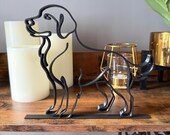 Front view of a modern Labrador or Golden Retriever line art sculpture, 3D printed in matte black from one flowing line. Features floppy ears, alert stance, and wagging tail on a flat black base. Displayed on a wood shelf with LED candles, gold candle holder, and greenery in gold pot. Ideal for dog-themed gifts, pet memorials, or stylish home decor for animal lovers.