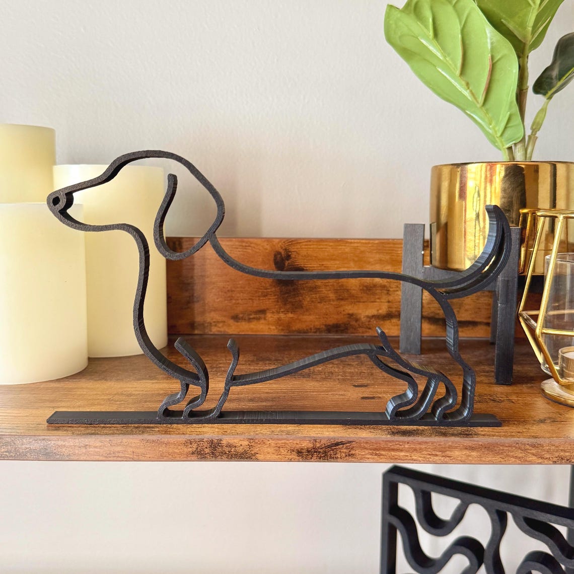 Minimalist dachshund sculpture in matte black displayed on a wooden shelf with candles and plants. Modern single line design makes it an ideal bookshelf accent or thoughtful dog lover gift.