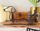 Minimalist dachshund sculpture in matte black displayed on a wooden shelf with candles and plants. Modern single line design makes it an ideal bookshelf accent or thoughtful dog lover gift.