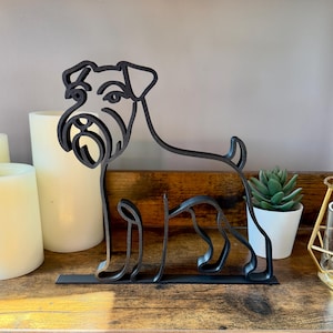 May include: A black metal sculpture of a dog, possibly a Schnauzer, in a minimalist style. The dog is standing on a flat base. The sculpture is on a wooden shelf with candles and a small potted plant in the background.