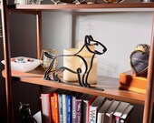 Styled lifestyle scene of a bull terrier line art sculpture on a wooden display shelf with vintage books, candles, glassware, and a curated bookshelf backdrop — modern minimalist dog home decor and a thoughtful bull terrier gift for dog moms, dog dads, and dog lovers.