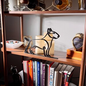 Styled lifestyle scene of a bull terrier line art sculpture on a wooden display shelf with vintage books, candles, glassware, and a curated bookshelf backdrop — modern minimalist dog home decor and a thoughtful bull terrier gift for dog moms, dog dads, and dog lovers.