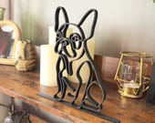Side-angled view of black French Bulldog line art sculpture on a rustic wood shelf with candles and gold candle holder behind it.