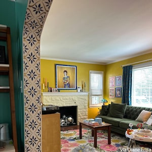 May include: A living room featuring mustard yellow walls, a fireplace with a white stone surround, and a green tufted sofa. A framed portrait of David Bowie hangs above the mantel. A floral rug covers the floor, and an arched doorway has a blue floral pattern.