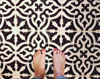 May include: A black and white tile stencil with a geometric pattern. The stencil is being used to create a floor design. The floor is painted white with black accents. The stencil is placed on the floor and the person is standing on the stencil. The text "Toledo Tile Stencil | Royal Design Studio" is visible in the bottom left corner of the image. The text "via Cara's Color Me Vintage" is visible in the bottom right corner of the image.