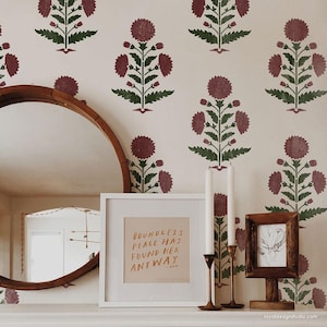 May include: A cream-colored wall with a repeating floral pattern in burgundy and green. A large, round, wooden-framed mirror hangs on the wall. A framed print with the text "BOUNDLESS PEACE HAS FOUND HER ANYWAY" sits on a shelf, along with candles and a small framed picture.