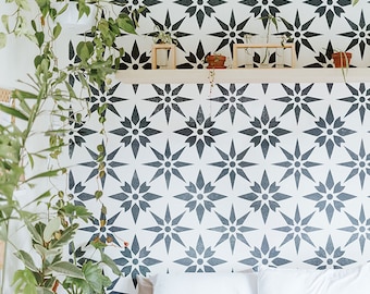 Puede incluir: Una pared blanca con una plantilla de estrella geométrica en blanco y negro. La plantilla se repite en toda la pared. Hay plantas colgando de la parte superior de la pared. Una cama blanca con almohadas blancas está frente a la pared.