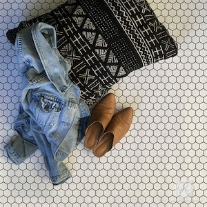 May include: A black and white patterned throw pillow with a denim jacket and two brown leather mules on a white hexagonal tile floor.