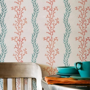 May include: Wallpaper with a repeating pattern of coral and leafy vines in teal and coral against a cream background. A teal teacup, saucer, and bowl are on a table with an orange napkin. A wooden chair is in the foreground.