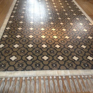 May include: A brown and white patterned floor stencil with a fringe border. The stencil is called "Chez Ali Moroccan Stencil" and is by "Royal Design Studio".