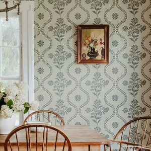 May include: A dining room with a light green wall featuring a repeating floral stencil pattern. The stencil design includes a daisy-like flower and a leafy vine. A framed painting of a floral arrangement hangs on the wall. Two wooden chairs are placed at a wooden table.