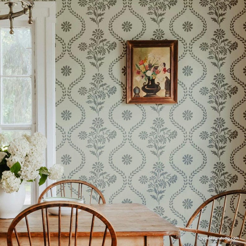 May include: A dining room with a light green wall featuring a repeating floral stencil pattern. The stencil design includes a daisy-like flower and a leafy vine. A framed painting of a floral arrangement hangs on the wall. Two wooden chairs are placed at a wooden table.