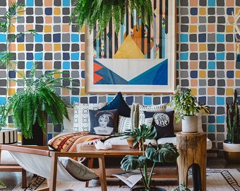 May include: A living room with a white sofa and a variety of plants. The walls are covered in a colourful geometric wallpaper with blue, yellow and grey squares. There is a framed abstract painting on the wall above the sofa.