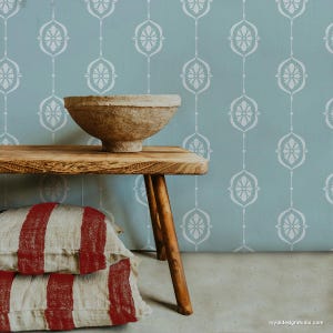 May include: A light blue wall with a white stencil pattern of repeating floral designs. A wooden bench with two red and white striped pillows sits in front of the wall. A brown bowl sits on top of the bench.