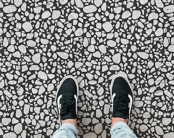 May include: A black and white patterned floor with a pebble design. Two black trainers are standing on the floor.