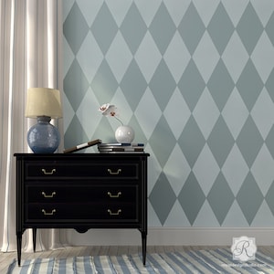 May include: A black three-drawer chest with brass handles and a blue lamp with a beige lampshade sits in front of a wall with a diamond pattern. A vase with a white flower and books are on top of the chest. A striped rug is on the floor.