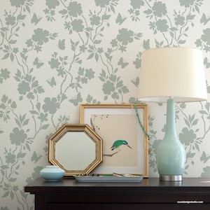 May include: A serene interior scene featuring a light blue lamp with a cream shade, a gold-framed mirror, and a framed bird print. The background is a floral wallpaper in light blue and white. A small blue vase and a decorative tray are also present.