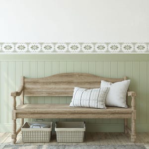 May include: A wooden bench with a curved backrest and turned legs, featuring two wire storage baskets underneath. Two throw pillows are placed on the seat: one white with thin black vertical stripes and one solid white. The wall has a stencil border.