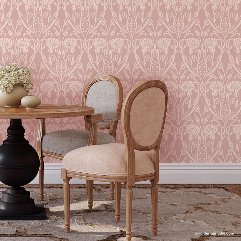 May include: Two wooden chairs with light pink upholstery sit at a wooden table in front of a pink wall with a white floral pattern.