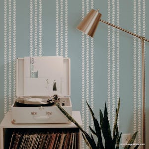 May include: A vintage record player with a white finish and a record on the turntable. The record player is sitting on a shelf with a stack of records underneath. A plant is in the foreground and a lamp is in the background. The wall behind the record player has a white and blue geometric pattern.